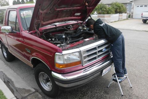 1994 Ford Bronco - Man inspects the engine bay of a red Ford Bronco, hood open, parked on a residential street.