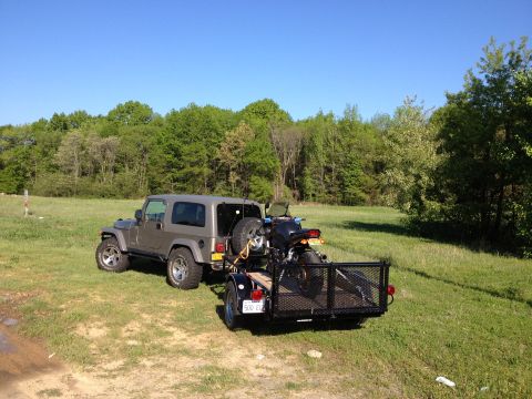 2005 Jeep Wrangler - A Jeep Wrangler transports a motorcycle on a trailer through a field on a clear day.