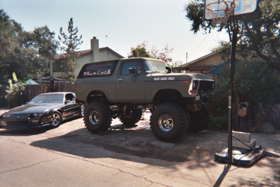 Lifted older Ford Bronco on large tires in driveway, black Camaro parked alongside, basketball hoop nearby