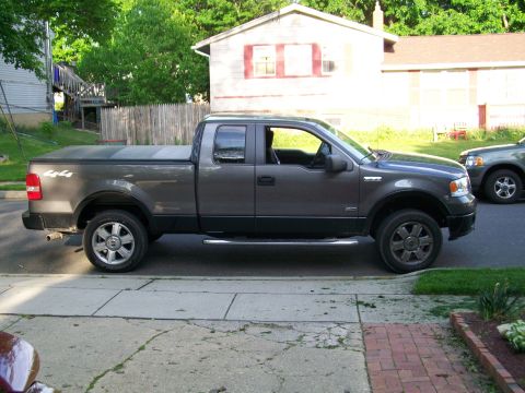 2008 Ford F-150 - gray Ford F-150 extended-cab 4x4 pickup with tonneau cover and 4.6 badge parked curbside by a pink house.