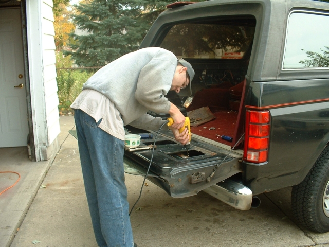 Person drilling into the dropped tailgate of a dark green Ford Bronco in a driveway