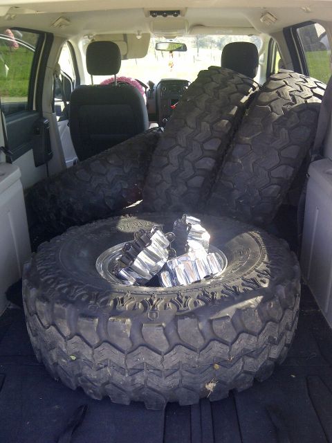 1995 Ford Bronco - The cargo area of a vehicle is filled with multiple off-road tires and a set of chrome lug nuts.