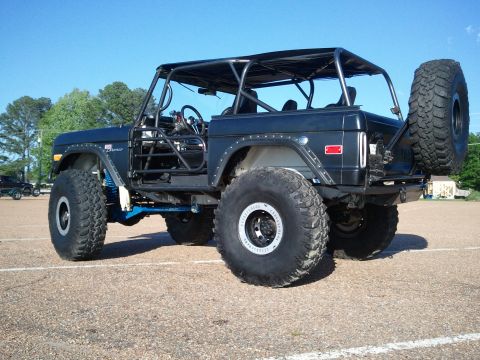 1967 Ford Mustang - Three-quarter rear view of a topless flat-black early Ford Bronco on a lift kit, showing an exposed roll cage, riveted fender flares, blue…