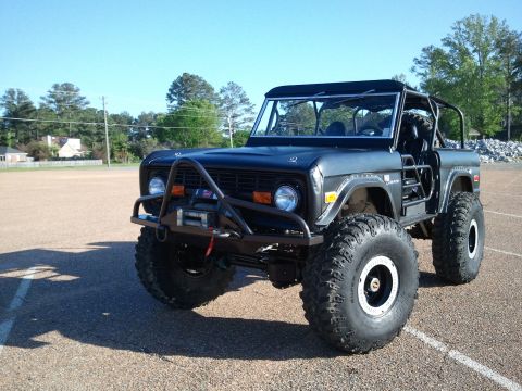 1967 Ford Mustang - Flat-black early Ford Bronco built as a topless trail rig, shot from the driver's front three-quarter on an empty asphalt lot. It runs a tu…