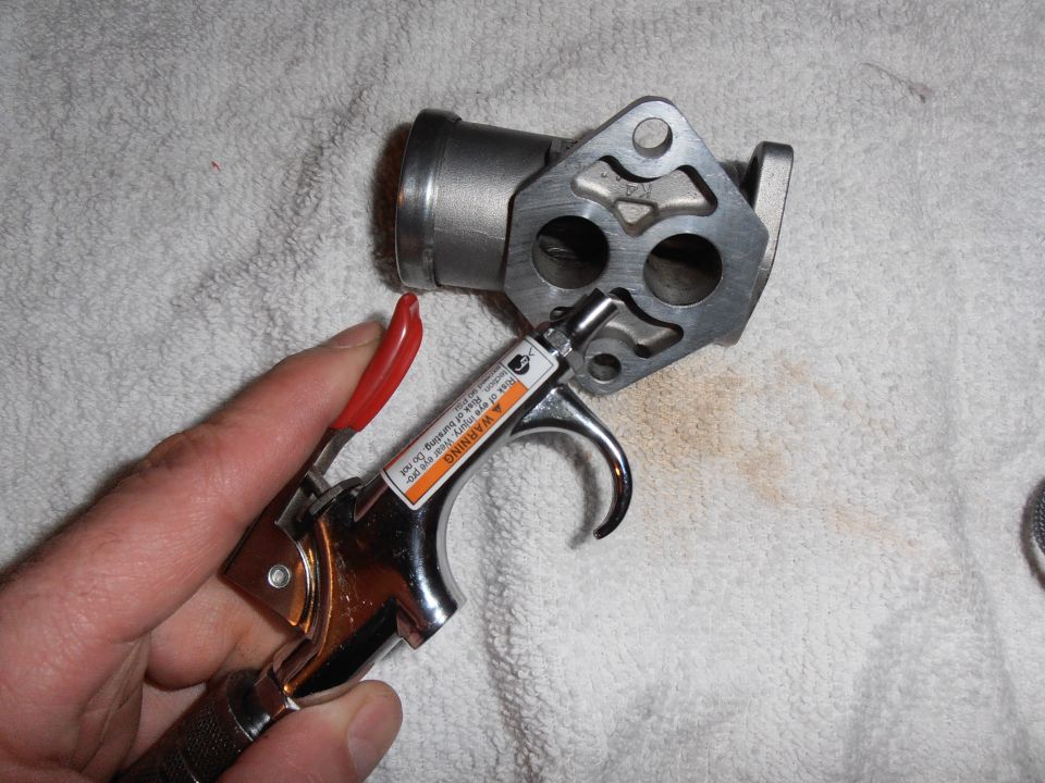 Hand holding an air duster next to a metal throttle body part. Warning label visible on the duster