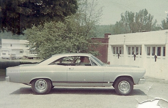 Side view of a silver 1966 Ford Fairlane hardtop parked on gravel in front of a white building