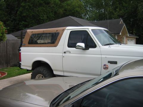 1992 Ford Bronco - Passenger-side view of a white Ford Bronco in a residential driveway with a tan soft top in place of the hardtop, a chrome mirror, whip ant…