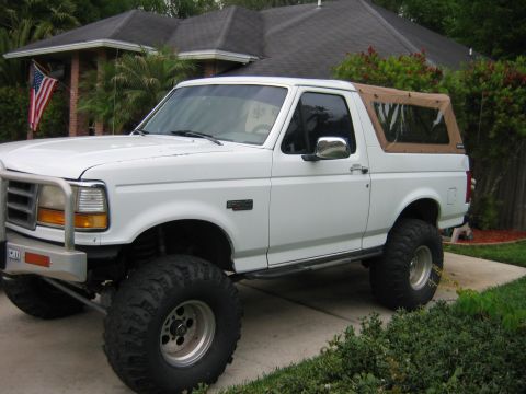 1992 Ford Bronco - A white Ford Bronco full-size SUV with a tan soft top, shown from a 3/4 front angle in a driveway. It has a lift kit, oversized mud tires,…