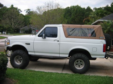 1992 Ford Bronco - white lifted 1990s full-size Ford Bronco fitted with a tan soft top and oversized tires, parked on a lawn in a tropical residential setting.