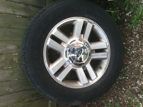 1993 Ford Crown Victoria - A single silver wheel with a chrome Ford logo is shown against a wooden fence and gravel background.