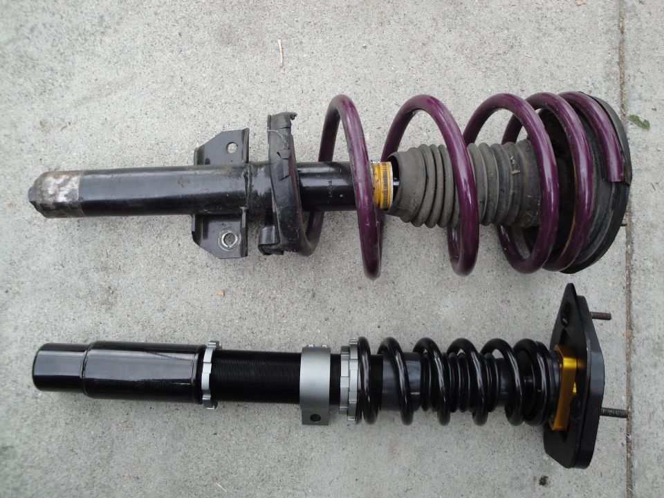 Two car suspension coilover assemblies, one with a purple spring and one with a black spring, laid on a concrete surface
