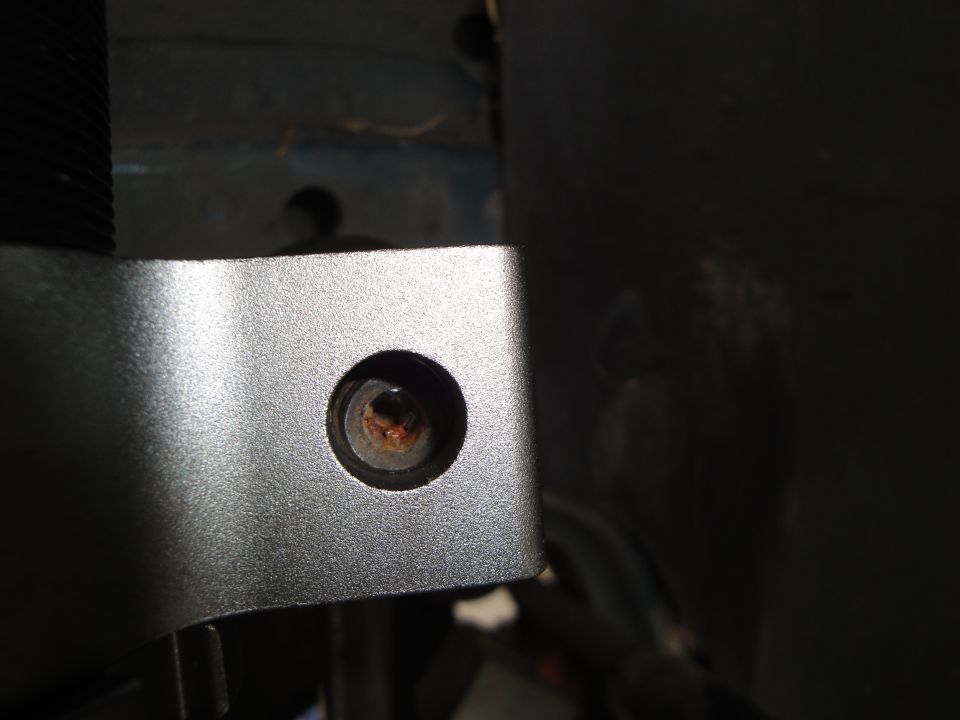 Close-up of a silver bracket with a hex bolt showing rust. Part of a black ribbed component is visible to the left