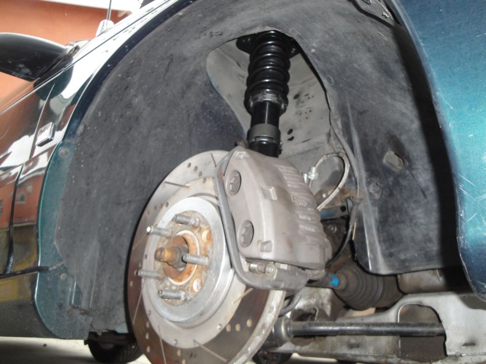 Close-up of a car's front wheel well showing a coilover shock, drilled brake rotor, and caliper