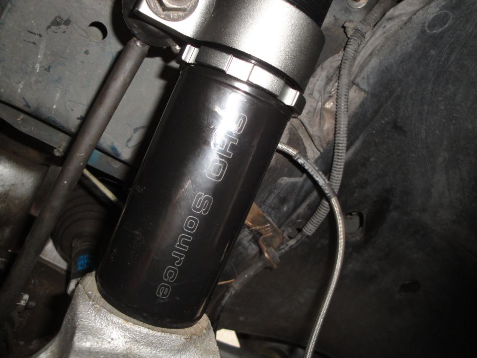 Close-up of a black shock absorber with "SHO SOURCE" etched on it, part of a car's suspension