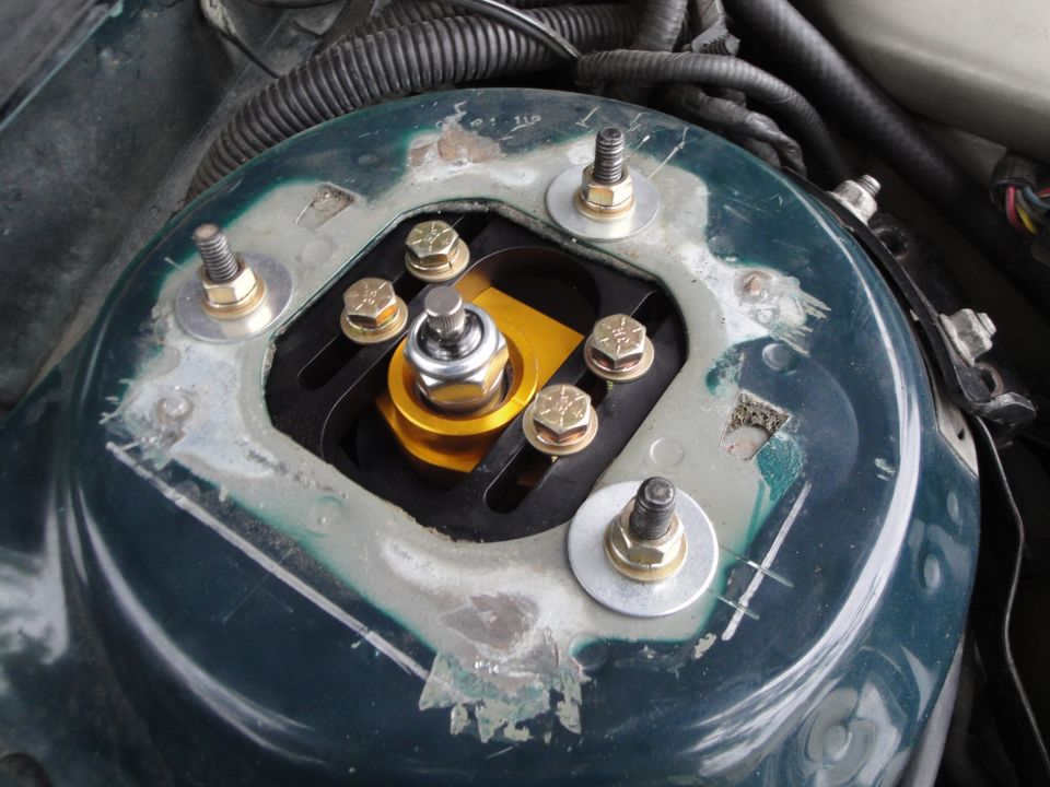 Green vehicle strut mount with aftermarket gold and black adjustable coilover assembly, showing bolts and washers