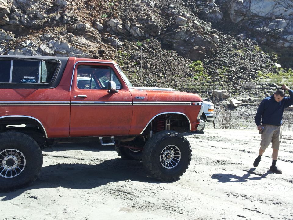 Orange 1978 Ford Bronco with big tires and lifted suspension parked on dirt. Another white truck is behind it. Guy stands to the right