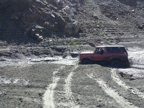 1978 Ford Bronco - A red 1978 Ford Bronco navigates a muddy, off-road terrain with steep hills in the background.