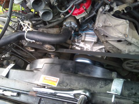 1994 Ford Bronco - Close-up view of the engine bay, focusing on the serpentine belt system and fan shroud. A 'Caution' sticker is present on the shroud.
