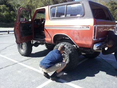 1978 Ford Bronco - Two individuals attend to an orange 1978 Ford Bronco, which has its driver's door ajar and is equipped with oversized tires.