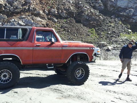 1978 Ford Bronco - A lifted 1978 Ford Bronco sits parked on a dirt lot, with a man standing to its right and another vehicle in the background.