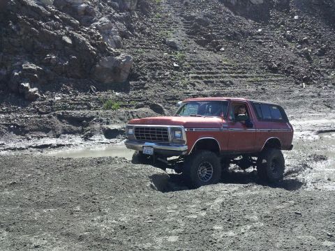 1978 Ford Bronco - Red 1978 Ford Bronco navigates a muddy, off-road terrain with a steep, rocky hillside in the background.