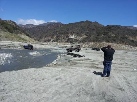 1978 Ford Bronco - Off-road vehicles traverse a rocky, dry riverbed under a clear blue sky with mountainous terrain.