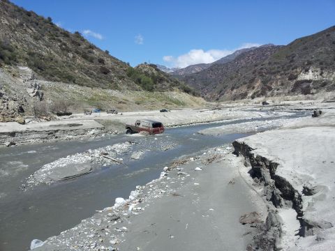 1978 Ford Bronco - Several vehicles navigate a dry riverbed surrounded by steep, rocky hills under a clear blue sky.