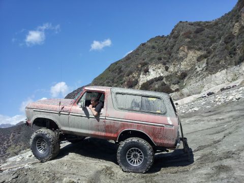 1978 Ford Bronco - Mud-covered 1978 Ford Bronco navigates a steep, rocky off-road path under a blue sky.