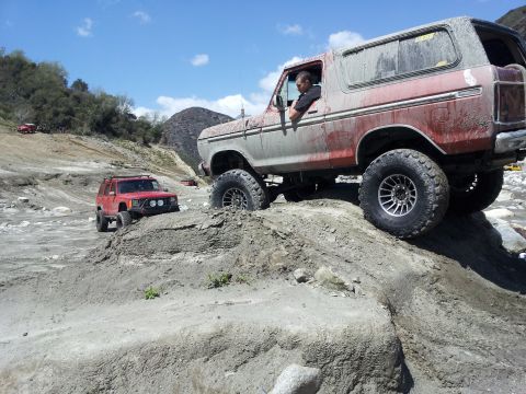 1978 Ford Bronco - Two off-road vehicles navigate a rocky, dry riverbed under a bright blue sky.