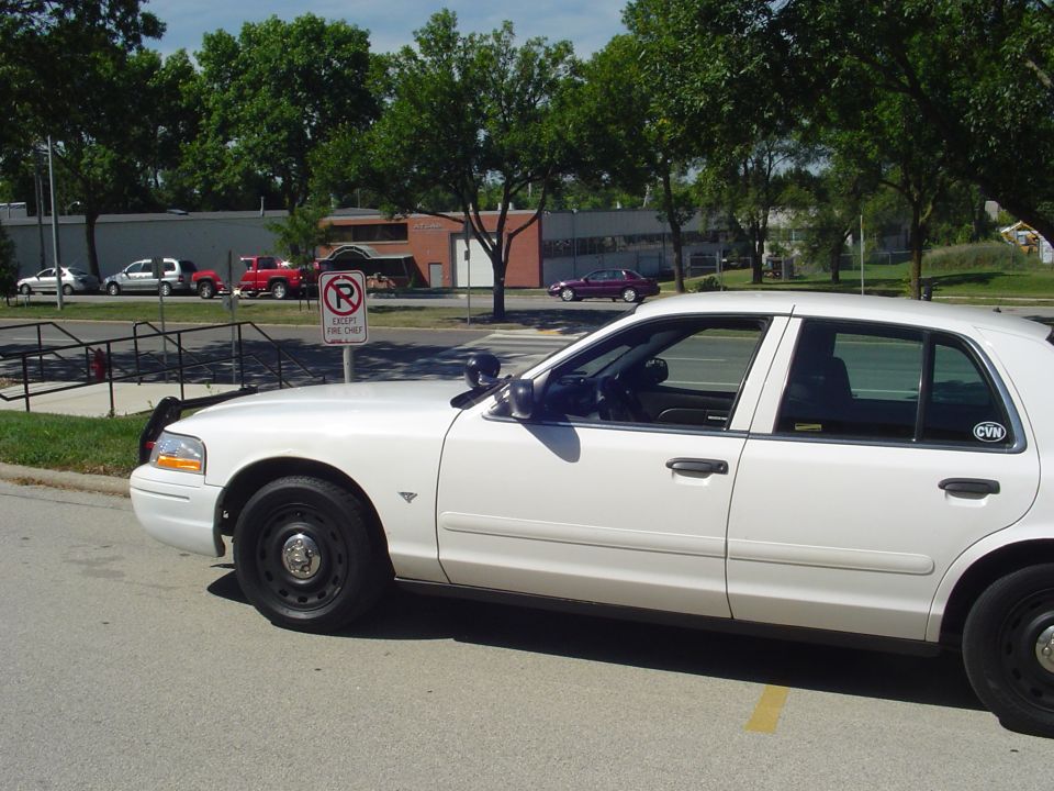 Side view of a white 2003 Ford Crown Victoria police car parked on asphalt. Other vehicles and trees are visible in the background