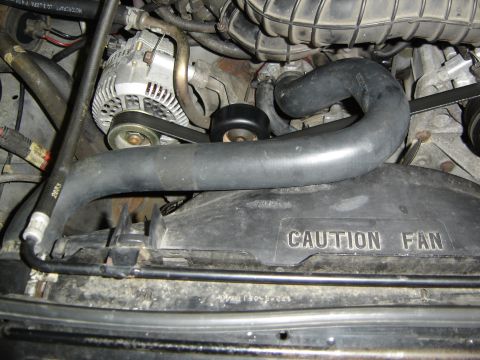 1996 Ford Bronco - Engine bay view over the radiator showing the black upper radiator hose arcing across a plastic fan shroud molded with "CAUTION FAN," with…