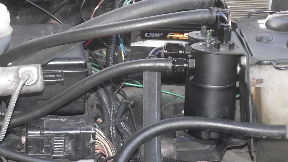 Black oil catch can with braided hoses in engine bay. Crane FireS logo visible