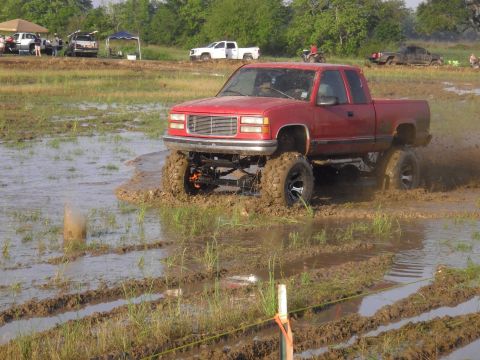 1993 Ford Bronco - A red pickup truck navigates a muddy track with tall tires, while other vehicles and tents are visible in the background.