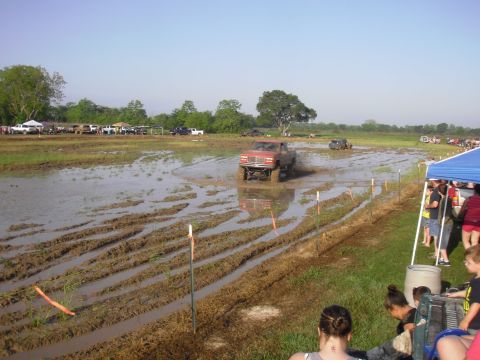 1993 Ford Bronco - A red pickup truck navigates a muddy track during an off-road event, with spectators gathered nearby.