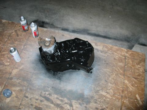 1984 Ford Bronco - An automotive transfer case is painted black with spray cans on a plywood surface.