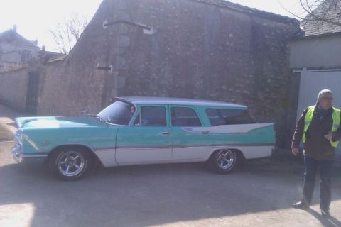 1991 Ford LTD Crown Victoria - A 1957 Plymouth Belvedere station wagon is parked outside a stone building, with a man in a yellow vest standing nearby.
