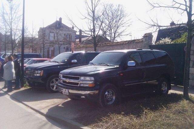Two black Chevrolet Suburbans parked side-by-side on a street. People stand near the front vehicle