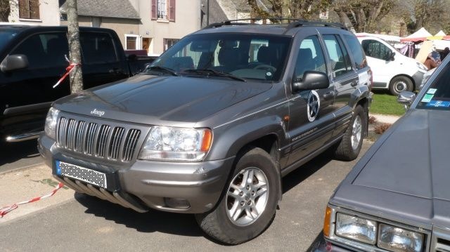 Gray Jeep Grand Cherokee parked next to a black pickup truck and a silver car. A white van is in the background