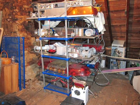 1991 Ford LTD Crown Victoria - A collection of emergency lights and electronic components are stored on shelving units in a cluttered attic space.