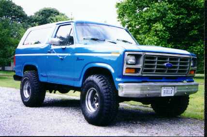 1985 Ford Bronco - This blue Ford Bronco is shown from a three-quarter front view, parked outdoors with trees behind it.