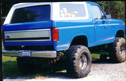 1985 Ford Bronco - Rear view of a lifted blue Ford Bronco with a white hardtop and off-road tires.