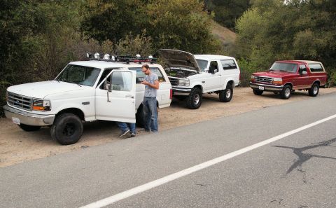 1994 Ford Bronco - Three Ford Broncos are parked on the shoulder of a road, with one white Bronco's hood open and a person standing by the driver's door.