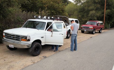 1994 Ford Bronco - Two people interact near a white Ford F-250 truck, with a red Ford Bronco parked behind it on a roadside.