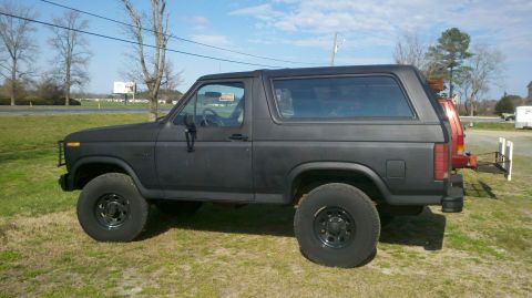 1986 Ford Bronco - This black Ford Bronco is seen from the side, parked outdoors on a grassy area under a partly cloudy sky.