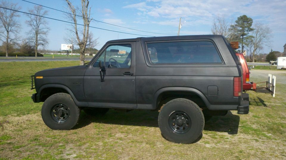 Black Ford Bronco parked on grass, side view. Another vehicle is partially visible behind it