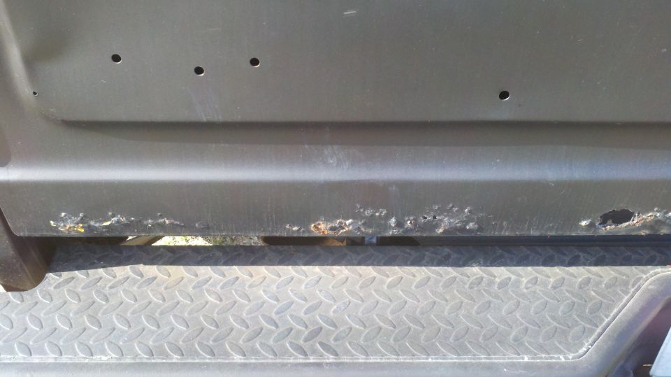Close-up of a dark-colored vehicle's lower body panel showing significant rust and several small holes