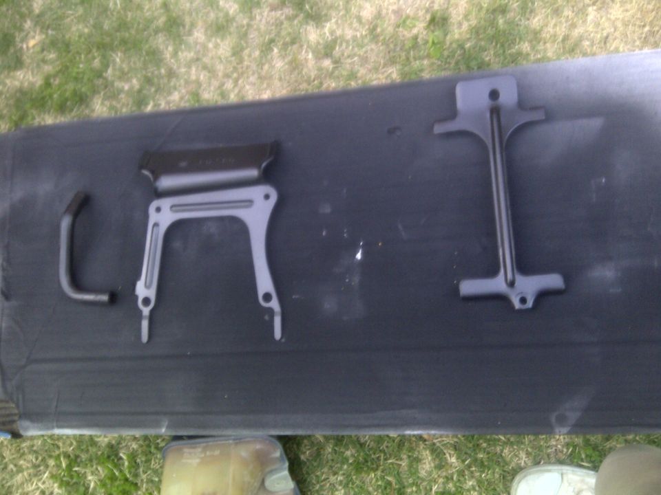 Three metal car parts, possibly brackets, laid out on a dark mat outdoors