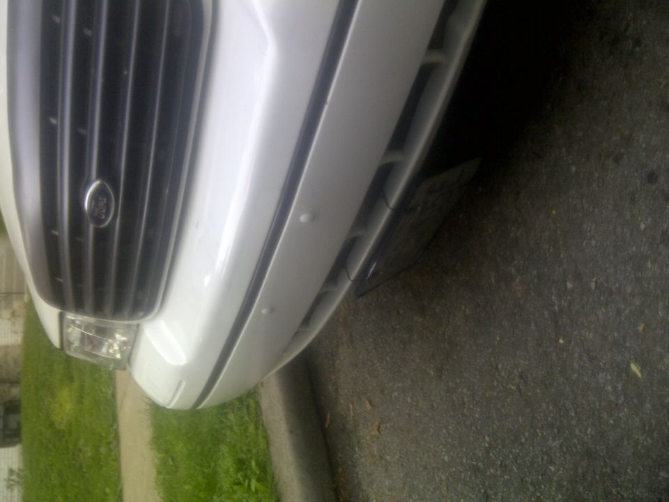 Close-up of a white car's front grille and bumper. The grille has vertical black bars and a Ford emblem