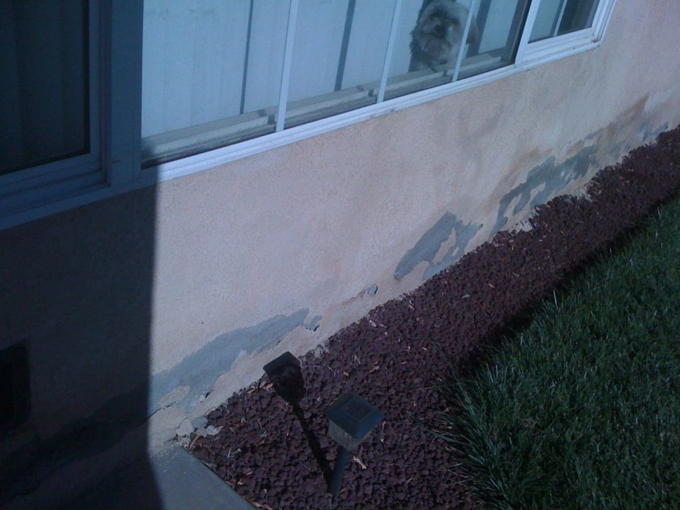 Building exterior wall with peeling stucco, dark red lava rock landscaping, and grass