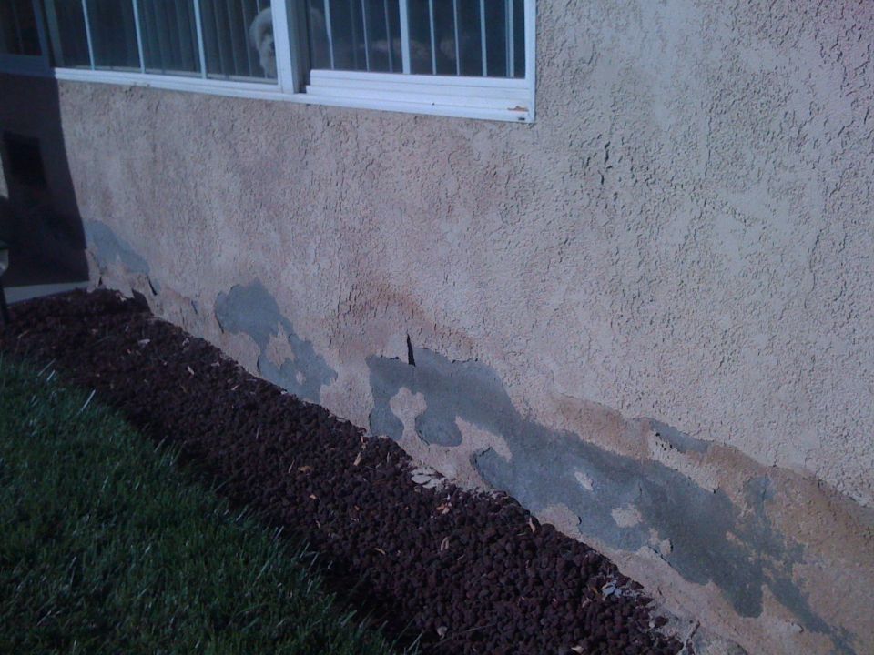 Damaged stucco wall of a building with a window and a strip of dark mulch next to grass